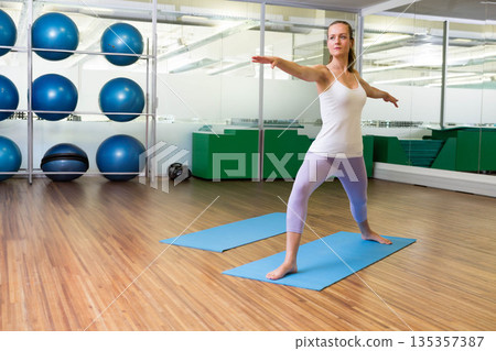 Woman holding yoga pose in fitness studio near blue mats and mirrored wall, copy space 135357387