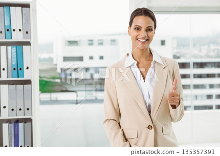 Businesswoman in suit giving thumbs-up in office by binder shelves with skyline view, copy space 135357391