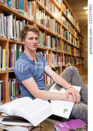 Male student sitting on carpeted library aisle studying open textbooks beside wooden bookshelf 135357402