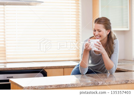Woman leaning on granite kitchen island holding ceramic mug under wooden blinds, copy space 135357403