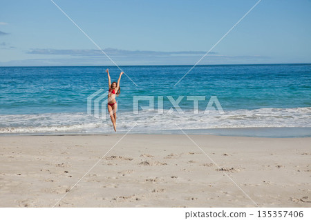 Woman jumping above shoreline on sandy beach by ocean wearing red two-piece swimsuit with blue sky Woman jumping above shoreline on sandy beach by ocean wearing red two-piece swimsuit with blue sky 135357406