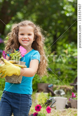 Girl holding concrete pot filled with pink petunias in sunny backyard garden wearing yellow gloves 135357407