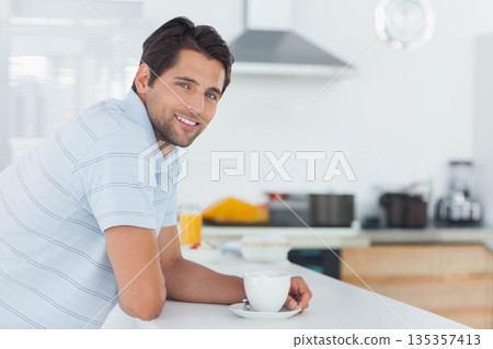 Man leaning against kitchen island holding coffee cup smiling beside orange juice, copy space Man leaning against kitchen island holding coffee cup smiling beside orange juice, copy space 135357413