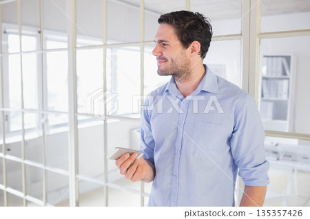 Man standing near glass partition in modern office wearing button-down shirt and holding smartphone 135357426
