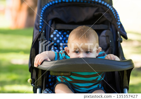 Infant boy gripping stroller tray bar and peeking over polka dot seat cushion in sunny park 135357427