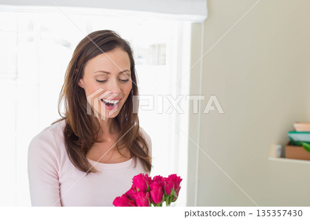 Woman holding pink roses bouquet by window beside shelf with green bowl and tan box 135357430