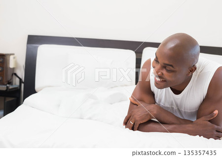 African American man smiling and reclining on bed wearing white tank top near telephone, copy space 135357435
