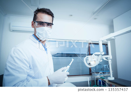 Male dentist wearing lab coat with mask, gloves holding polishing tool in clinic, copy space 135357437