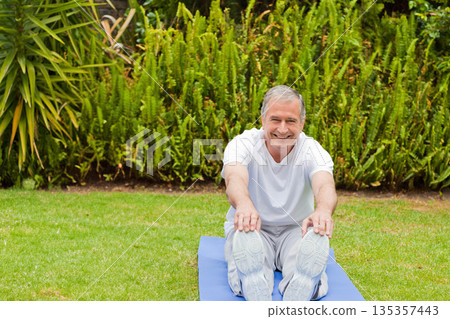 Senior man in sportswear stretching forward on blue exercise mat in backyard garden 135357443