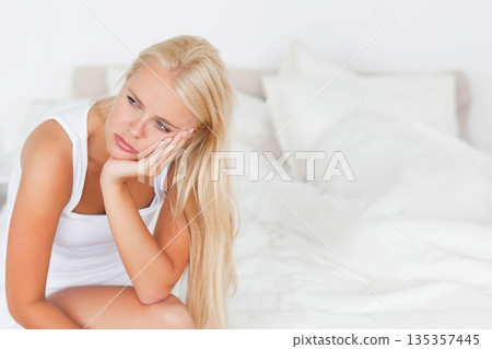 Woman leaning forward resting face on hand gazing right on bed with white pillows, copy space 135357445