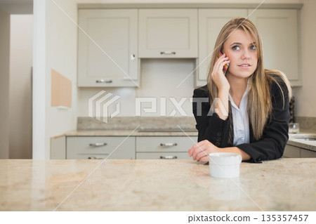 Female in blazer talking on smartphone near white mug while leaning on kitchen counter, copy space Female in blazer talking on smartphone near white mug while leaning on kitchen counter, copy space 135357457