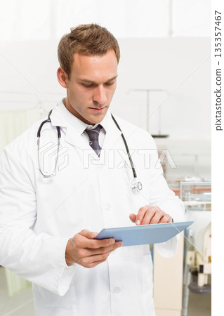 Male doctor wearing lab coat with stethoscope reviewing data on light blue tablet in exam room Male doctor wearing lab coat with stethoscope reviewing data on light blue tablet in exam room 135357467
