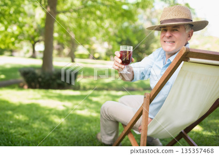 Senior man sitting in deckchair on lawn wearing straw hat holding stemmed wine glass, copy space Senior man sitting in deckchair on lawn wearing straw hat holding stemmed wine glass, copy space 135357468