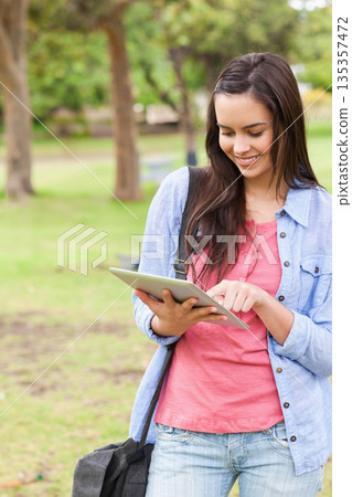 Woman holding tablet and smiling while standing in grassy park with shoulder bag, copy space 135357472