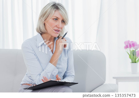 Mature female consultant sitting on grey sofa at office holding clipboard and pen, copy space Mature female consultant sitting on grey sofa at office holding clipboard and pen, copy space 135357473