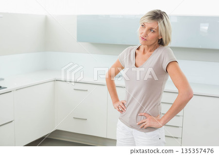 Blonde woman standing in home kitchen gazing at white countertop and gray backsplash, copy space Blonde woman standing in home kitchen gazing at white countertop and gray backsplash, copy space 135357476