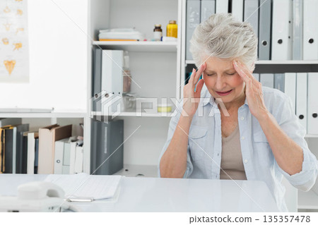 Senior woman pressing temples with hands at clinic desk with telephone and notebook, copy space Senior woman pressing temples with hands at clinic desk with telephone and notebook, copy space 135357478