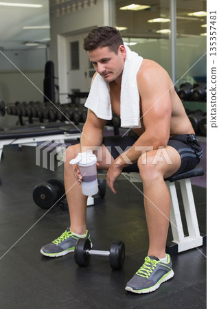 Male fitness enthusiast sitting on weight bench with towel drinking from shaker bottle in gym 135357481