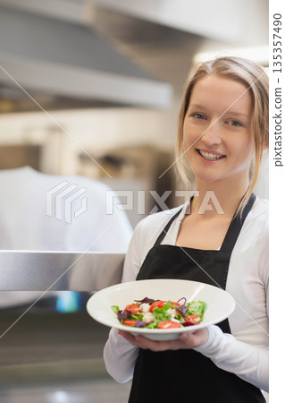 female chef standing under commercial oven hood by pass in kitchen holding salad plate, copy space 135357490