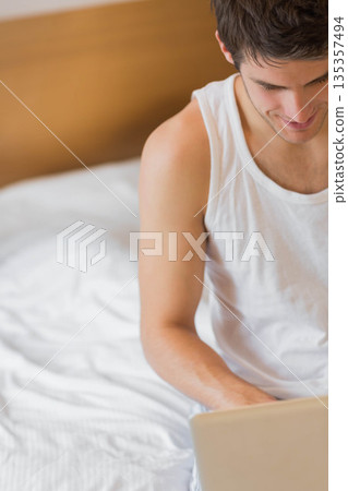 Man sitting on bed in bedroom using laptop amid white sheets, wooden headboard, copy space Man sitting on bed in bedroom using laptop amid white sheets, wooden headboard, copy space 135357494