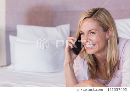 Middle-aged woman talking on smartphone while reclining on white pillows and headboard in bedroom 135357495