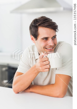 Man leaning on white kitchen counter and holding white ceramic coffee mug while gazing right Man leaning on white kitchen counter and holding white ceramic coffee mug while gazing right 135357496