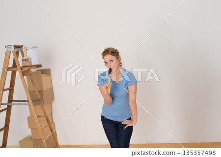 Female standing in empty room holding paintbrush to lips, near ladder with paint bucket and boxes 135357498