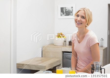 Blonde woman smiling while sitting at kitchen counter with orange juice glasses and coffee carafe 135357501