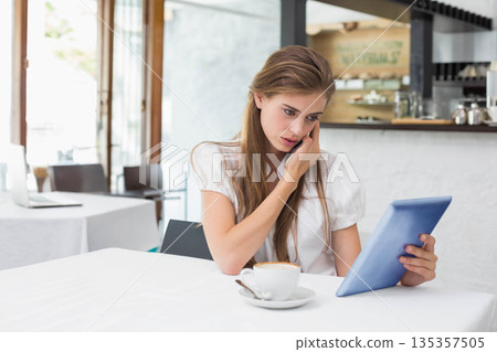 Woman in her twenties talking on smartphone and holding tablet at cafe table with coffee cup 135357505