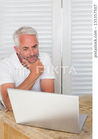 Senior man wearing white polo sitting at desk using silver laptop in home office, copy space 135357507