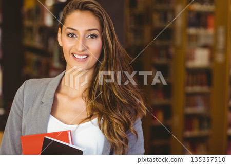 Woman wearing grey blazer standing inside library holding red notebook and black folder smiling Woman wearing grey blazer standing inside library holding red notebook and black folder smiling 135357510