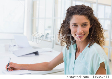 Woman holding pen over notepad smiling at camera at office desk with laptop beside glass partition Woman holding pen over notepad smiling at camera at office desk with laptop beside glass partition 135357511