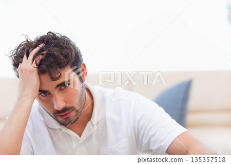 Man sitting on beige couch by overexposed window in room running hand through hair with cushion 135357518