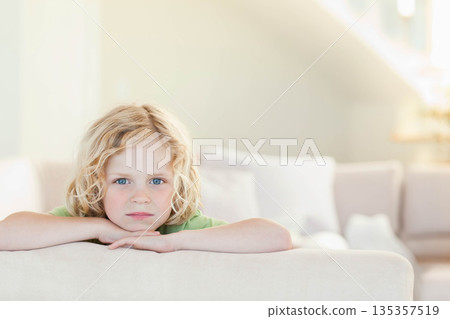 Boy wearing green t-shirt leaning over back of sofa in living room near decorative cushions 135357519
