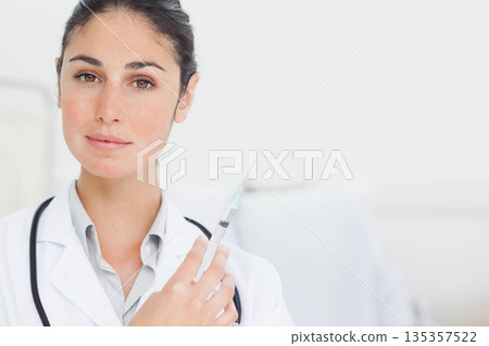 Female doctor holding syringe and wearing stethoscope and lab coat inside exam room, copy space 135357522