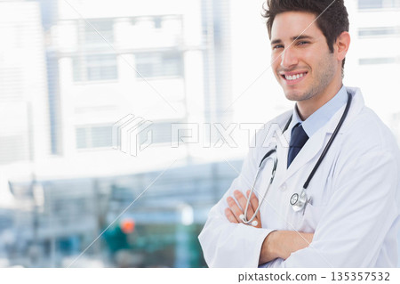 Male doctor smiling near windows in clinic reception wearing lab coat and stethoscope, copy space Male doctor smiling near windows in clinic reception wearing lab coat and stethoscope, copy space 135357532