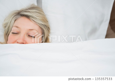 Mature woman lying under white duvet in bedroom with bed frame, white sheets and pillow 135357533