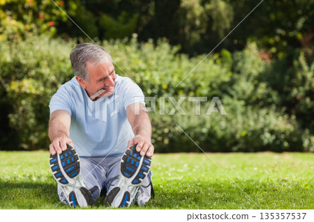 Senior man stretching legs on green lawn in park wearing sports T-shirt and running shoes Senior man stretching legs on green lawn in park wearing sports T-shirt and running shoes 135357537