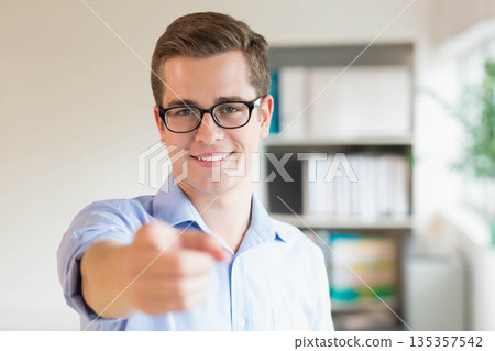 Man wearing light blue shirt and eyeglasses, pointing at camera in office by bookshelf and plant Man wearing light blue shirt and eyeglasses, pointing at camera in office by bookshelf and plant 135357542