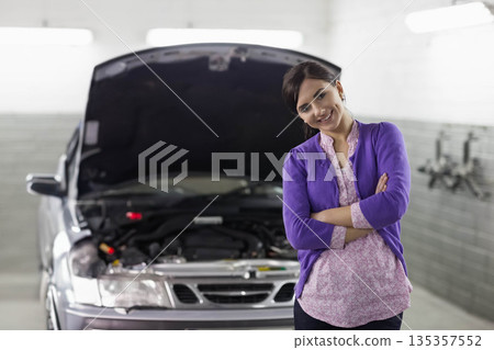 Woman standing arms crossed in service bay near silver sedan with open hood, copy space 135357552