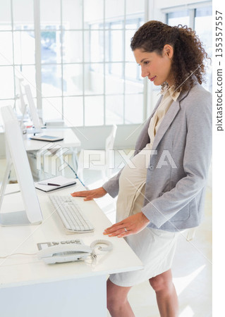 Pregnant woman working at standing desk in office using monitor, keyboard, phone, copy space Pregnant woman working at standing desk in office using monitor, keyboard, phone, copy space 135357557