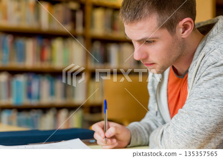 Male student writing in notebook at wooden table in library study with blue pen, copy space 135357565