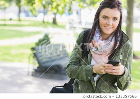 Woman wearing camouflage jacket, holding smartphone, carrying bag in sunlit park, copy space 135357566