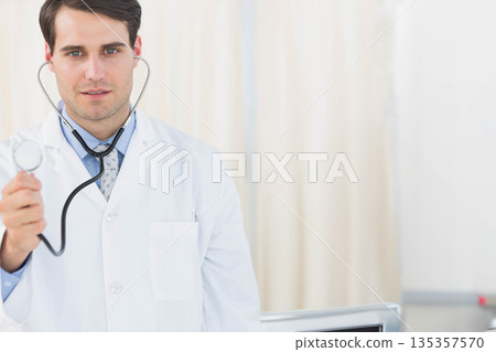 Male doctor wearing white coat holding stethoscope toward viewer in exam room with medical monitor Male doctor wearing white coat holding stethoscope toward viewer in exam room with medical monitor 135357570