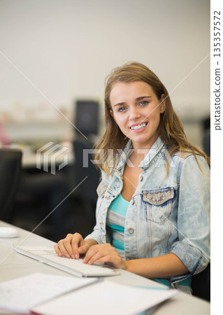 Female worker wearing denim jacket typing on keyboard at computer lab desk with monitor and papers 135357572
