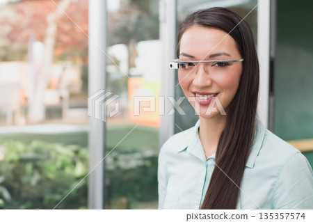 Asian woman standing inside modern office lobby using smart glasses near large glass panels Asian woman standing inside modern office lobby using smart glasses near large glass panels 135357574