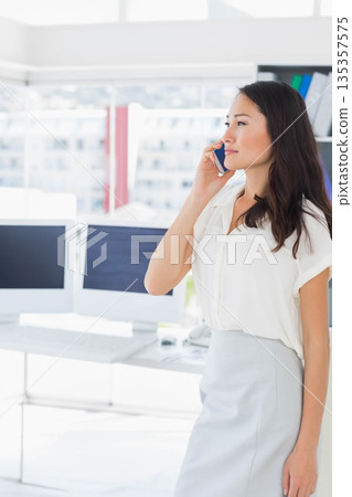Asian female professional talking on smartphone beside dual-monitor desk at office, copy space 135357575