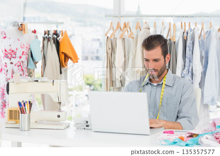 Male fashion designer sitting at studio desk by window working on laptop with sewing machine Male fashion designer sitting at studio desk by window working on laptop with sewing machine 135357577