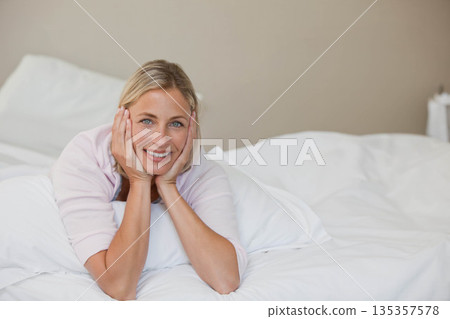 Smiling mid adult female lying on white bedding wearing light pink top hugging pillow in bedroom Smiling mid adult female lying on white bedding wearing light pink top hugging pillow in bedroom 135357578