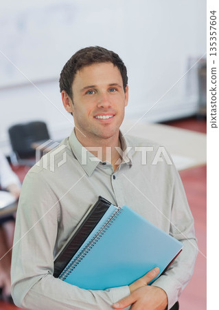 Man holding blue spiral notebook and black binder in classroom with desks chairs whiteboard Man holding blue spiral notebook and black binder in classroom with desks chairs whiteboard 135357604